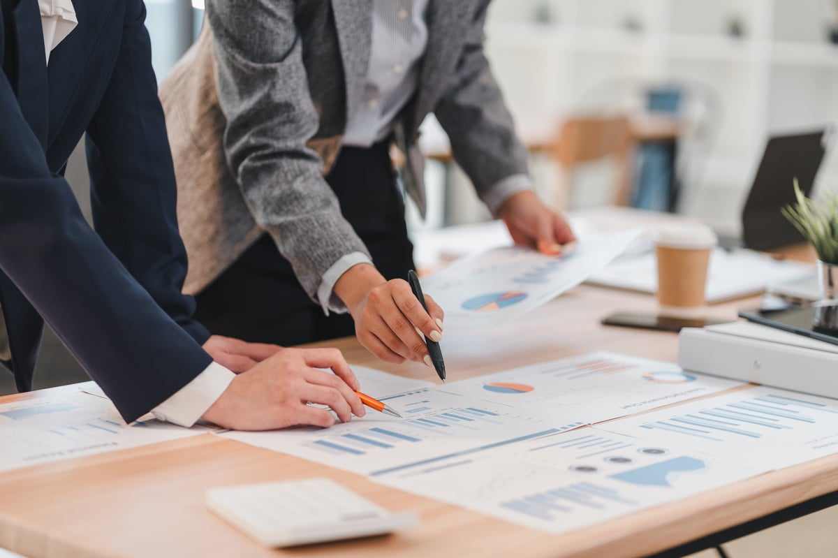 Two business professionals analyzing financial charts and graphs on wooden desk in modern office setting, collaborating on project with focus and teamwork