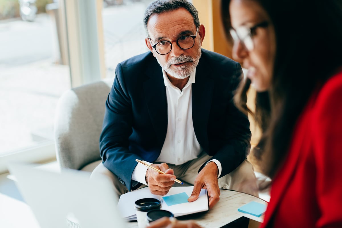 Focused senior manager conversing with a colleague in an office, noting important details during a management planning session