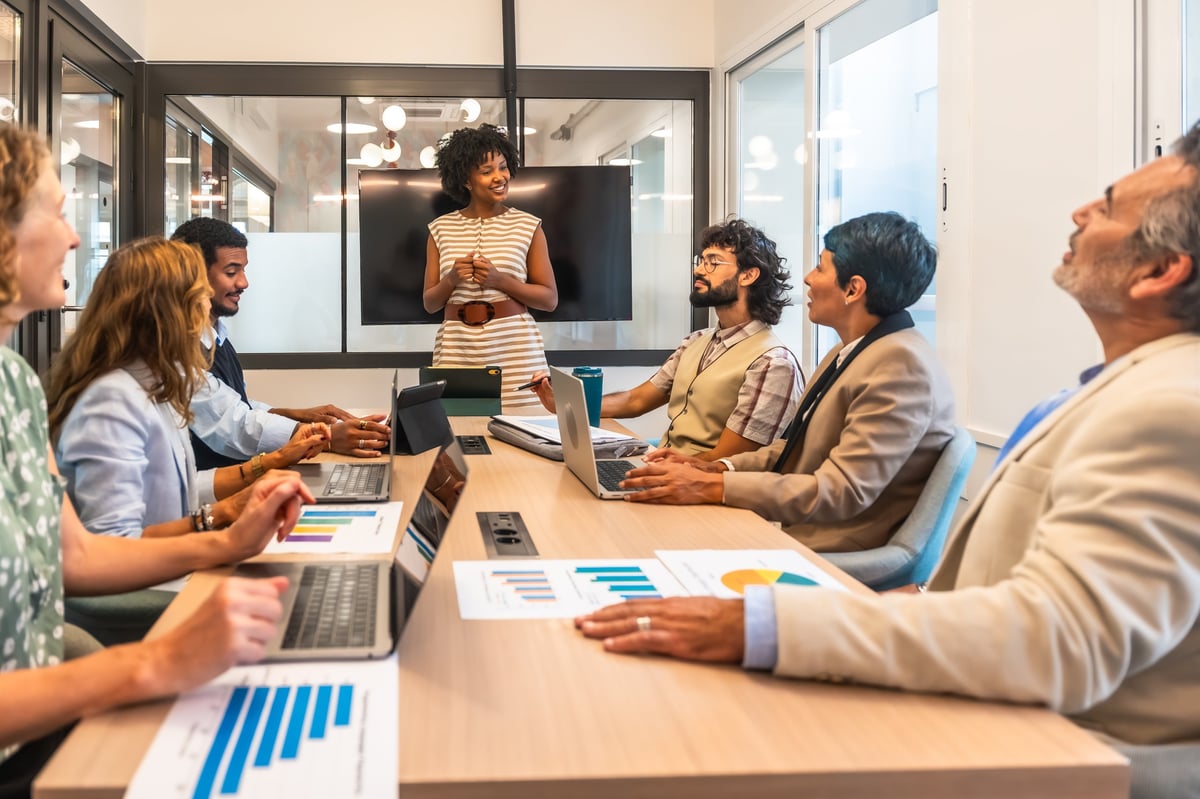 Diverse team of six professionals collaborating in a modern office meeting, woman presenting data and ideas to colleagues using laptop, tablet, charts and paperwork