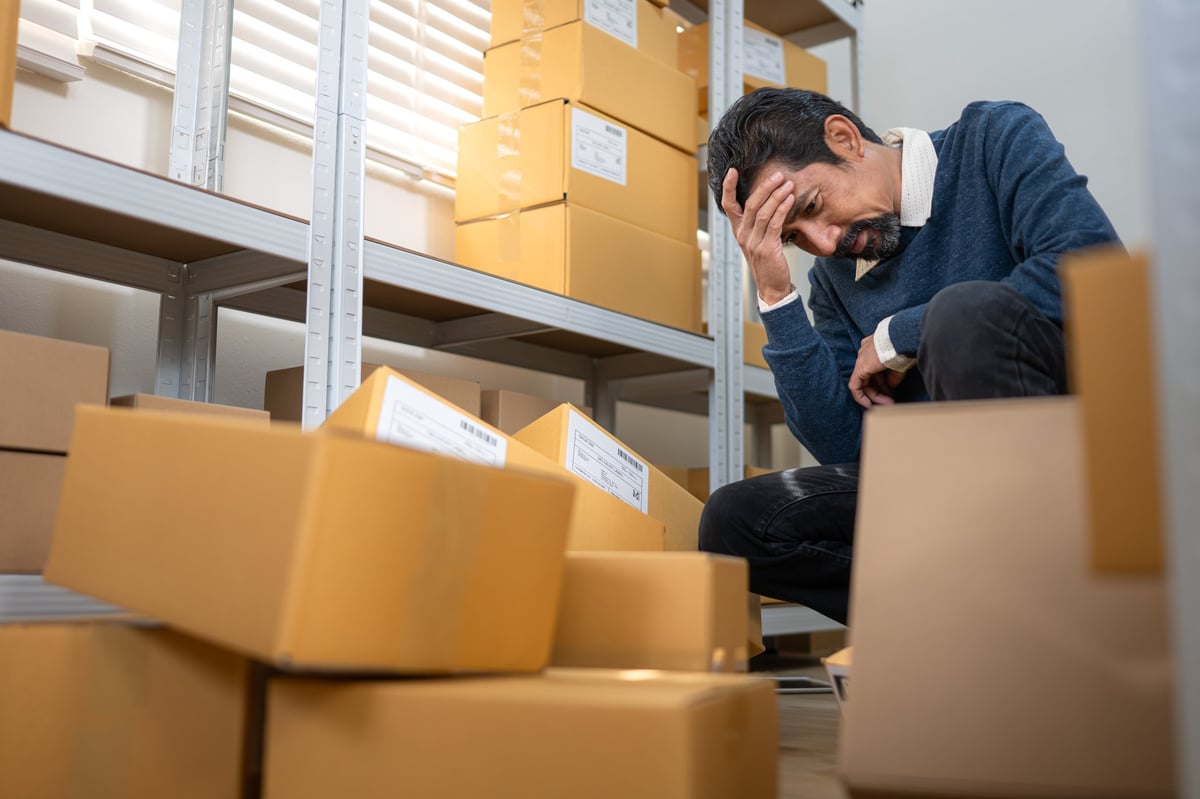 Stressed warehouse worker sitting among cardboard boxes, expressing business problem, inventory loss, and work pressure in storage room