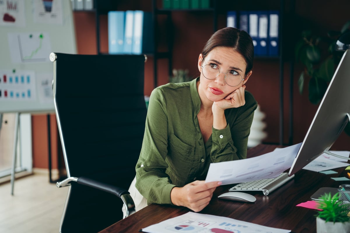 A young businesswoman in an office pauses at her desk, holding printed reports and studying charts with a thoughtful, concerned expression while working on financial analysis and paperwork.