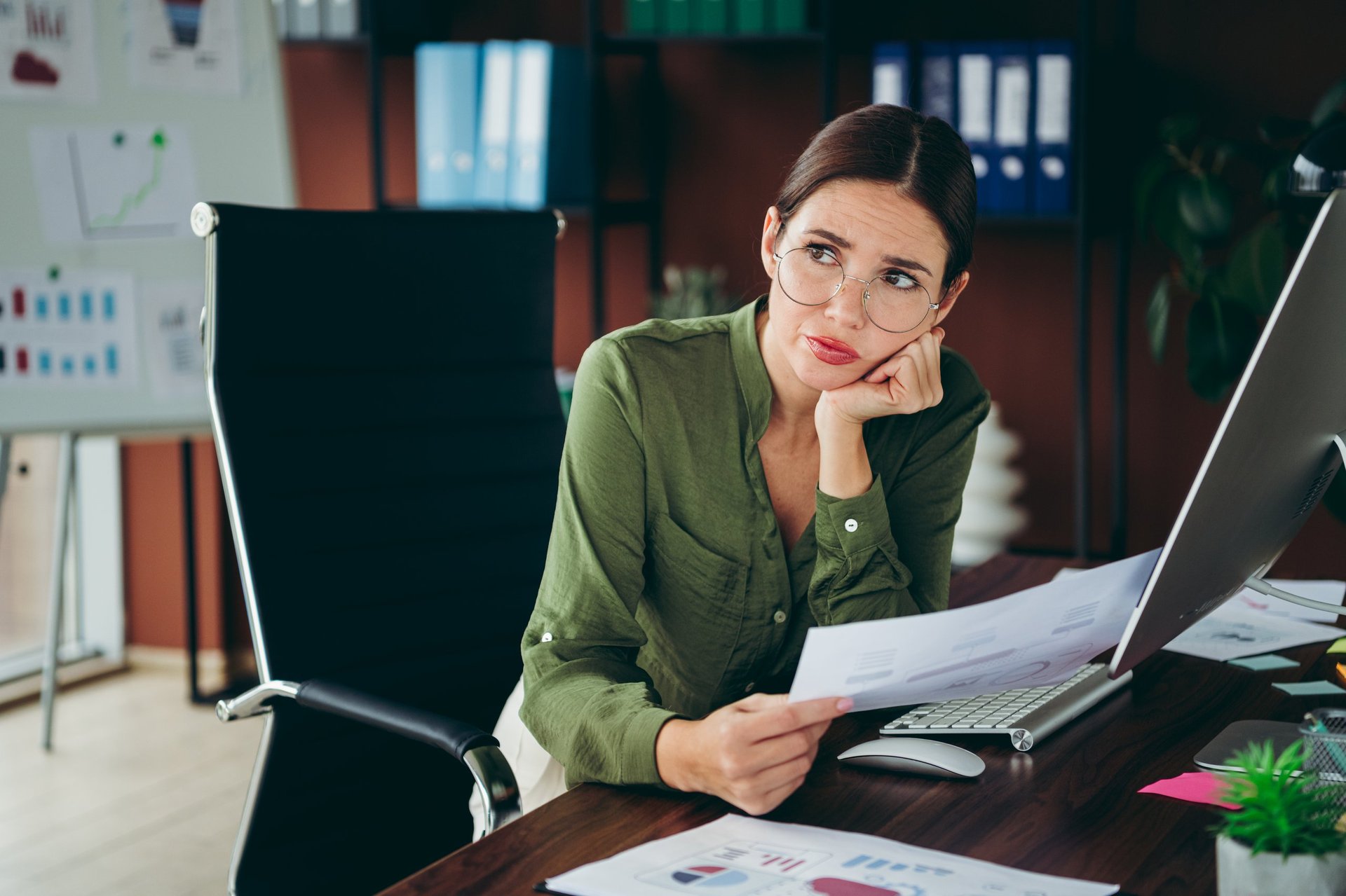 A young businesswoman in an office pauses at her desk, holding printed reports and studying charts with a thoughtful, concerned expression while working on financial analysis and paperwork.