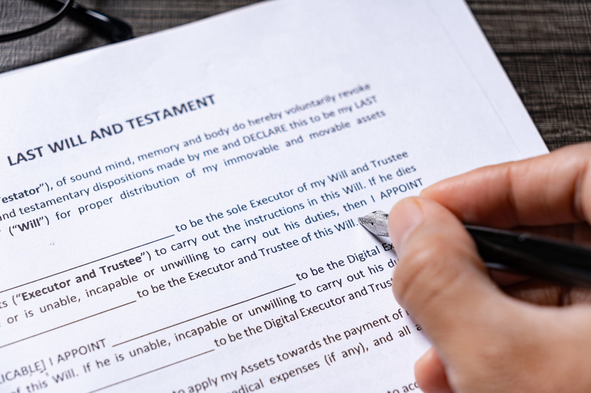 A detailed close up shot showing a hand holding a black fountain pen, touching the nib to a printed Last Will and Testament document on a wooden desk surface.