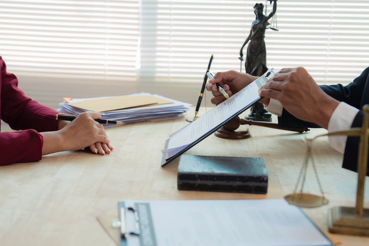 Close up of justice scales and judge gavel on wooden desk with cash bundle and lawyer consulting client about financial lawsuit in blurred background.